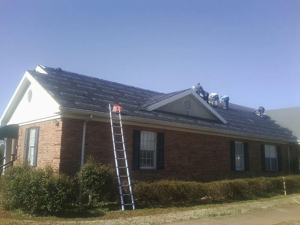 Crew tearing shingles off a brick ranch roof with a ladder set — Metal Roof Repair work by our crew in Watford City