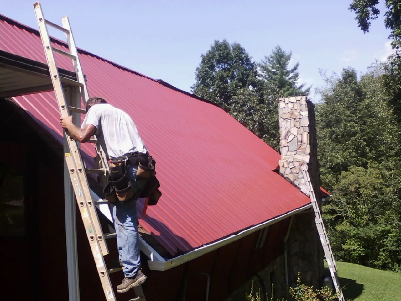 Roofer on a ladder beside a red metal roof and stone chimney — Soffit & Fascia Repair by Roofing Brothers in Watford City